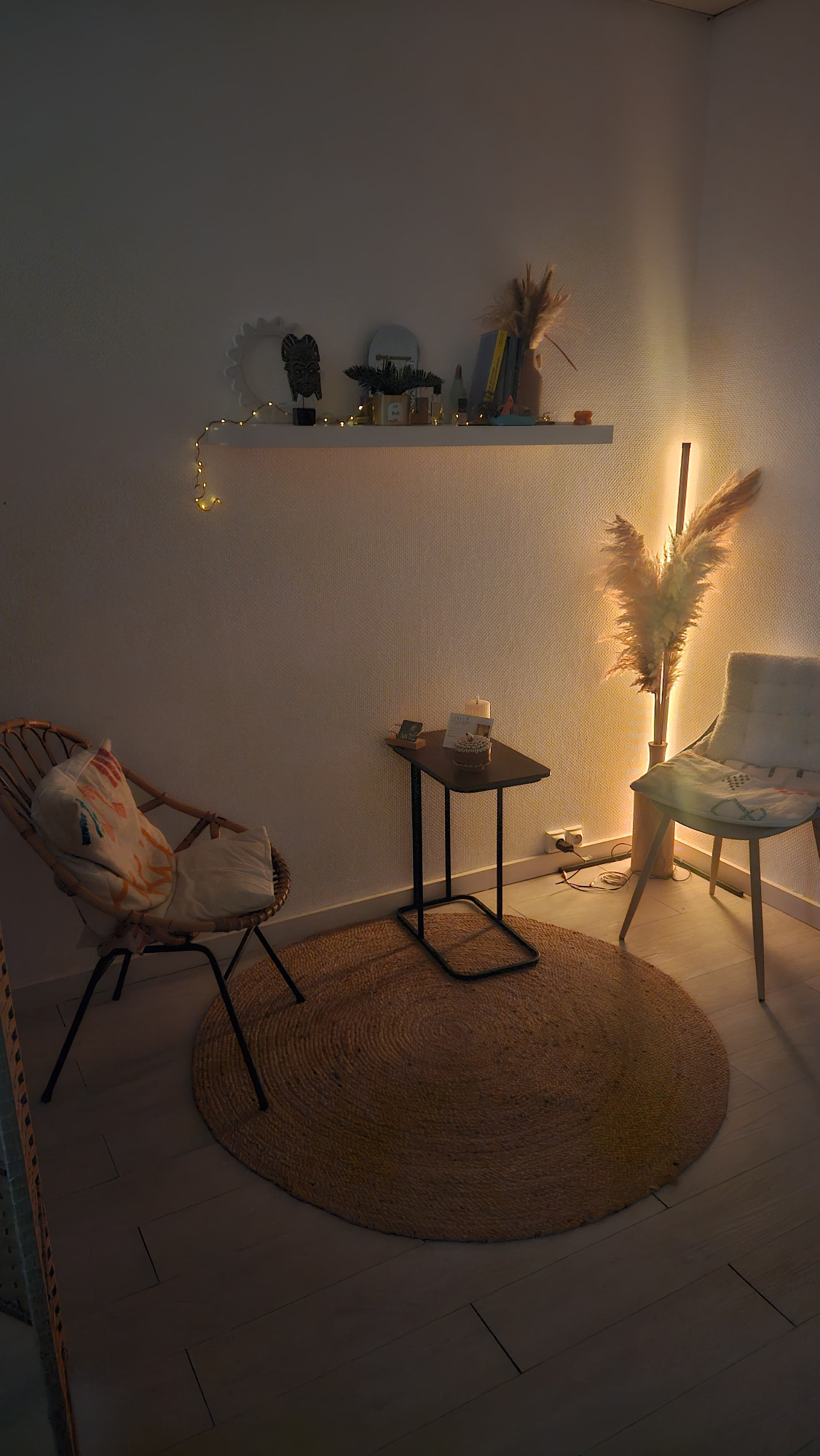 Salon de massage avec fauteuil en rotin, tapis rond en jute, table d’appoint, étagère décorative et éclairage d’ambiance chaleureux.